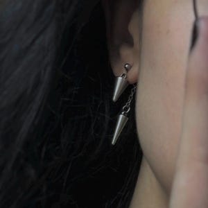 May include: A pair of silver cone-shaped earrings with a dangling chain and a small bead accent.