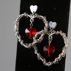 May include: Silver heart-shaped earrings with dangling red crystal hearts. The earrings are made of wire that is twisted into a heart shape.