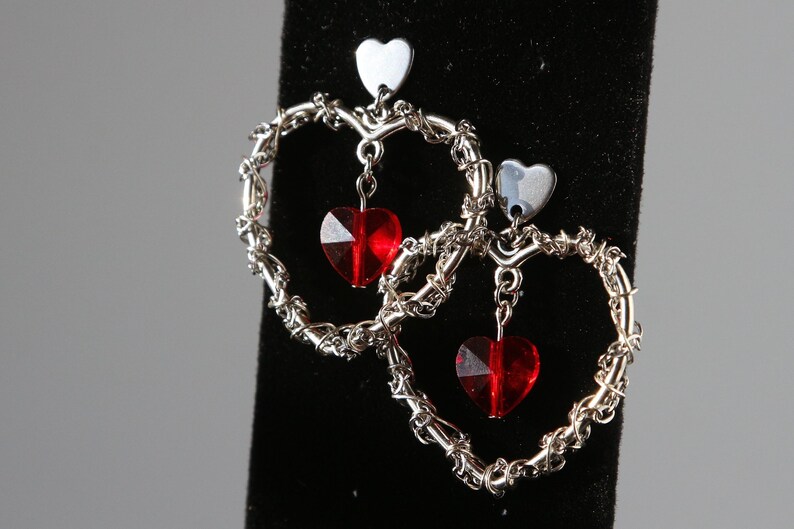 May include: Silver heart-shaped earrings with dangling red crystal hearts. The earrings are made of wire that is twisted into a heart shape.