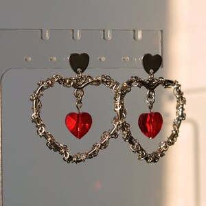 May include: A pair of silver heart-shaped earrings with dangling red heart-shaped crystals.