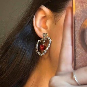 May include: Silver heart-shaped dangle earrings with a red crystal heart charm. The earrings have a delicate chain design.