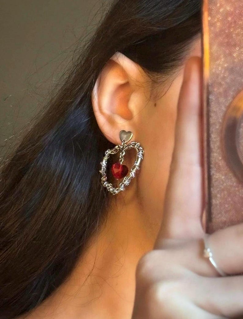 May include: Silver heart-shaped dangle earrings with a red crystal heart charm. The earrings have a delicate chain design.