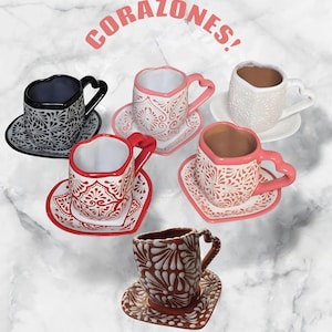 Puede incluir: Tazas de té y platillos de cerámica en forma de corazón en varios colores, incluyendo negro, blanco, rojo y rosa. Cada taza y platillo presenta un patrón floral decorativo. La palabra "CORAZONES!" está escrita arriba.