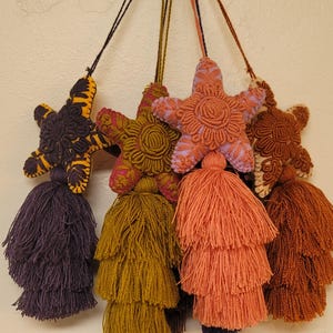 May include: Five star-shaped fabric ornaments with tassels. The stars are embroidered with different patterns in shades of brown, purple, and orange. The tassels are layered and come in shades of purple, brown, and orange.