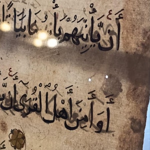 Puede incluir: Primer plano de un pergamino envejecido con caligrafía árabe negra. El texto está escrito en una escritura fluida, con trazos de tinta visibles y algunos elementos decorativos. El pergamino tiene un tono cálido y envejecido.