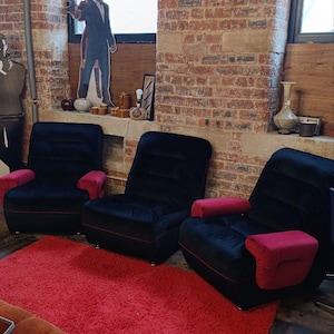 May include: Three black armchairs with red armrests and trim sit on a red shag rug. The chairs are in front of a brick wall with a window. A cardboard cutout of a man and woman is in the window.