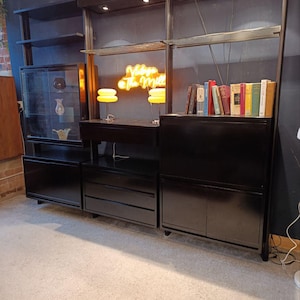 May include: A black modular shelving unit with integrated lighting, drawers, and cabinets. The unit features glass-fronted display cases, a desk area, and shelves holding books. A neon sign reads "Vintage The Mill".