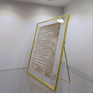 May include: A gold-colored metal frame with a glass front displays a vintage document with Arabic calligraphy. The frame is on a clear glass surface and is angled to the right.