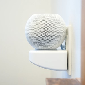 May include: A white smart speaker with a mesh grille is mounted on a white wall mount. The speaker is plugged into an electrical outlet.