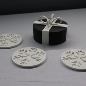 May include: Four white snowflake-shaped coasters with a black gift box with a white ribbon and bow on top.
