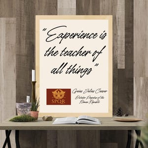 Puede incluir: Un cuadro enmarcado con la cita "La experiencia es el maestro de todas las cosas" de Cayo Julio César. El cuadro está sobre una mesa con una vela, una planta y un libro abierto. El fondo es una pared de madera.