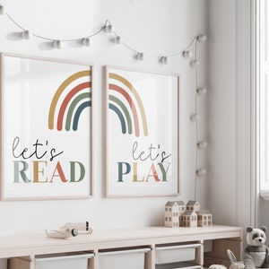 May include: Two framed prints with rainbow designs and the text "let's read" and "let's play" in colorful letters. The prints are hung on a white wall with a string of white lights.