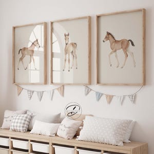 May include: Three framed watercolour prints of foals in a nursery. Each print shows a different pose of a light brown foal against a beige background. The frames are light wood, and the room is decorated with pillows and a banner.