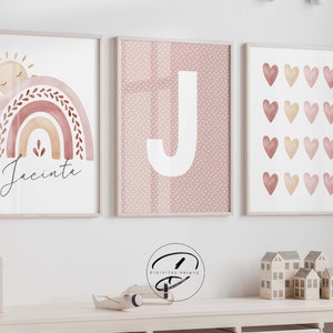 May include: Three framed prints in a pink and white colour scheme. The first print features a watercolour rainbow with a sun and the name Jacinta. The second print features a white letter J on a pink polka dot background. The third print features a grid of pink and orange watercolour hearts.