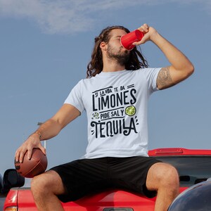 May include: A white t-shirt with the text "Si la vida te da limones, pide sal y tequila" is worn by a person sitting on a red truck. The person is drinking from a red cup and holding a football. A black grill is in the foreground.