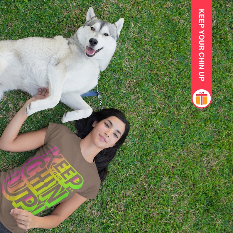 May include: A woman wearing a brown t-shirt with the text "Keep Your Chin Up" in rainbow colors, lying on her back in a grassy field with a husky dog lying on top of her.