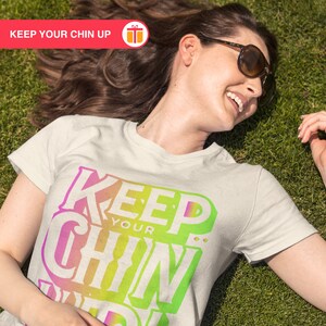 May include: A white t-shirt with a colorful graphic that says "Keep Your Chin Up". The shirt is being worn by a person lying on a green grassy surface.