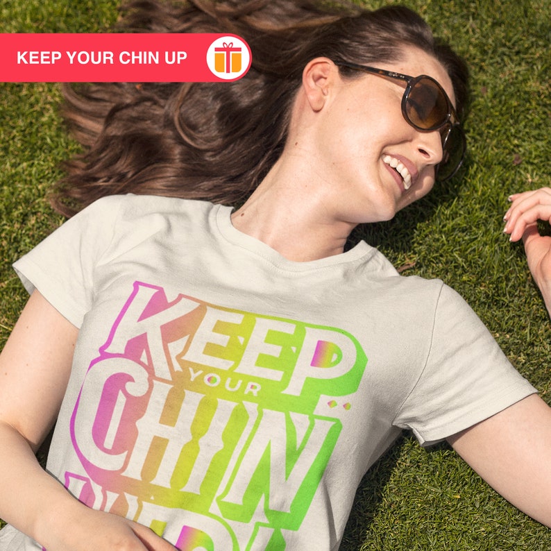 May include: A white t-shirt with a colorful graphic that says "Keep Your Chin Up". The shirt is being worn by a person lying on a green grassy surface.