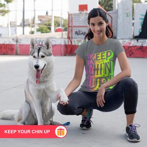 May include: A woman wearing a gray t-shirt with the text "KEEP YOUR CHIN UP" in colorful letters, is squatting down with a husky dog. The dog is wearing a collar and leash.