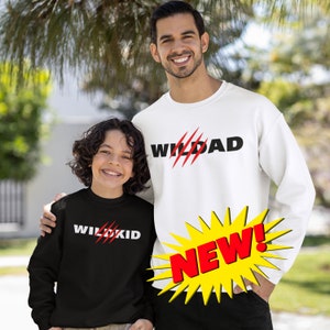 May include: A father and son wearing matching black and white sweatshirts. The father's sweatshirt says "Wildad" in black with red claw marks. The son's sweatshirt says "Wildkid" in white with red claw marks. The father is wearing a white sweatshirt and the son is wearing a black sweatshirt. The image has a yellow starburst with the word "NEW!" in red.