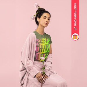 May include: A woman wearing a green t-shirt with the text "KEEP YOUR CHIN UP" in a rainbow gradient. She is wearing a pink cardigan and a pink skirt. She has flowers in her hair.