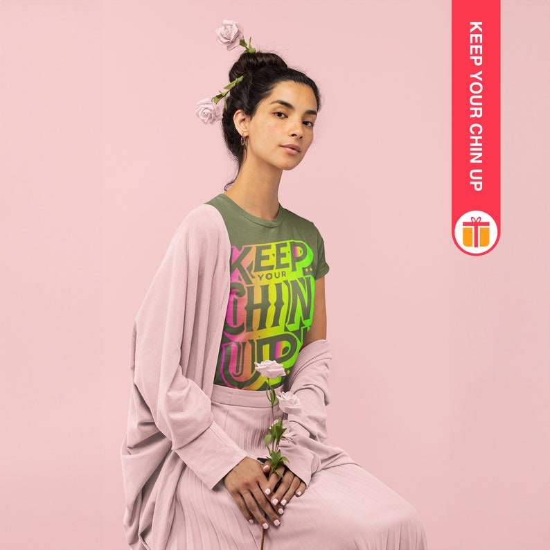 May include: A woman wearing a green t-shirt with the text "KEEP YOUR CHIN UP" in a rainbow gradient. She is wearing a pink cardigan and a pink skirt. She has flowers in her hair.