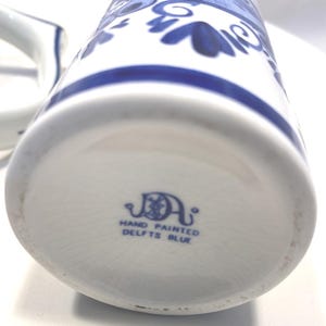 Op de afbeelding: Witte keramische kan met blauwe bloemmotieven en een blauwe streep. De onderkant van de kan heeft de tekst "HAND PAINTED DELFTS BLUE". Een klassiek stuk Delfts blauw.