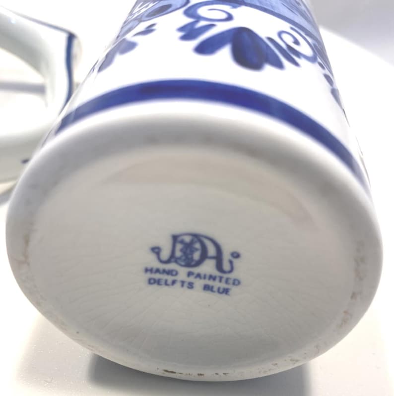 Op de afbeelding: Witte keramische kan met blauwe bloemmotieven en een blauwe streep. De onderkant van de kan heeft de tekst "HAND PAINTED DELFTS BLUE". Een klassiek stuk Delfts blauw.