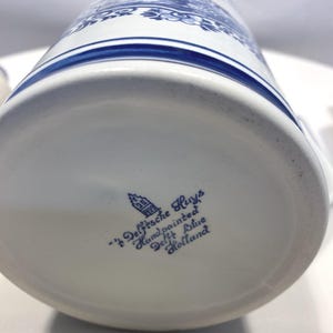 Op de afbeelding: Een witte keramische mok met blauwe bloemen- en architecturale ontwerpen. De onderkant van de mok heeft de tekst "'t Delftsche Huys Handpainted Delft Blue Holland" in blauwe letters.