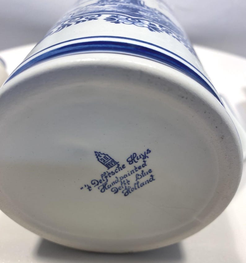 Op de afbeelding: Een witte keramische mok met blauwe bloemen- en architecturale ontwerpen. De onderkant van de mok heeft de tekst "'t Delftsche Huys Handpainted Delft Blue Holland" in blauwe letters.