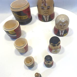 Könnte beinhalten: Eine Sammlung von Holz-Matrjoschka-Puppen, jede bemalt mit einem anderen Gesicht und Outfit. Einige Puppen sind zylindrisch, mit dem Wort "PRESIDENT" sichtbar. Die Puppen sind auf einer weißen Oberfläche angeordnet.