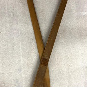 Könnte beinhalten: Ein Paar lange, braune Küchenzange aus Holz mit gekreuztem Design. Jeder Arm der Zange ist aus einem einzigen Stück Holz gefertigt, wobei ein Arm in einem länglichen ovalen Ausschnitt und der andere in einem runden Loch zum Greifen endet.