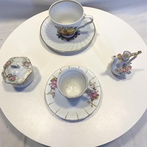 May include: A collection of vintage porcelain teacups, saucers, and a small sugar bowl with a lid. The teacups and saucers feature floral and fruit designs with gold trim. A small decorative creamer with pink roses is also present.