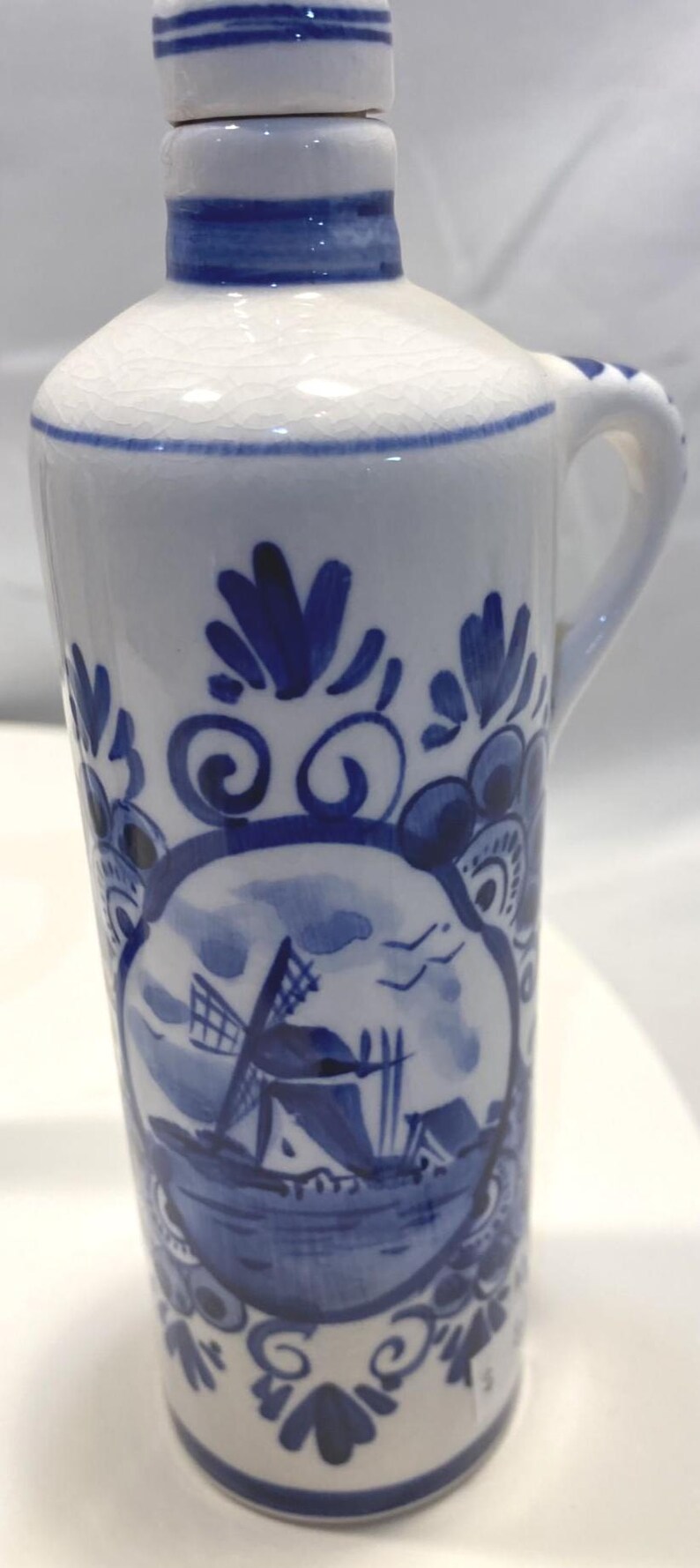 Op de afbeelding: Een witte keramische kan met blauwe bloemen- en windmolenontwerpen. De kan heeft een handvat en een tuit. Het ontwerp omvat een windmolen, een boot en gestileerde bloemen. De kan is ongeveer 15 cm hoog.