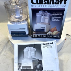 Op de afbeelding: Een Cuisinart Mini-Prep keukenmachine, met een doorzichtige plastic kom en witte basis, wordt getoond naast de doos en de handleiding. De doos toont de productnaam en vermeldt kenmerken zoals "Compact Powerhouse" en "Chops Grinds Purees".