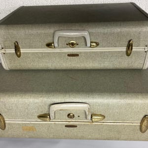 May include: Two vintage gray suitcases stacked on top of each other. The suitcases have white handles, brass-colored clasps, and a keyhole lock. The top suitcase is smaller than the bottom one.