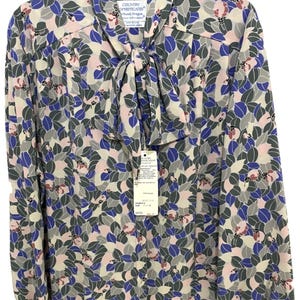 Puede incluir: Una blusa de manga larga con cuello de lazo y un patrón de hojas repetitivo en tonos azul, gris y rosa pálido sobre un fondo crema. La prenda incluye una etiqueta de marca que dice Country Sophisticates y una etiqueta de venta colgante.