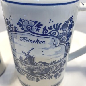 Op de afbeelding: Witte keramische mok met blauw bloemen- en windmolenontwerp. De mok heeft het woord "Heineken" in blauw schrift. Het handvat is wit en de rand heeft een blauwe streep. De mok heeft een klassieke, vintage esthetiek.