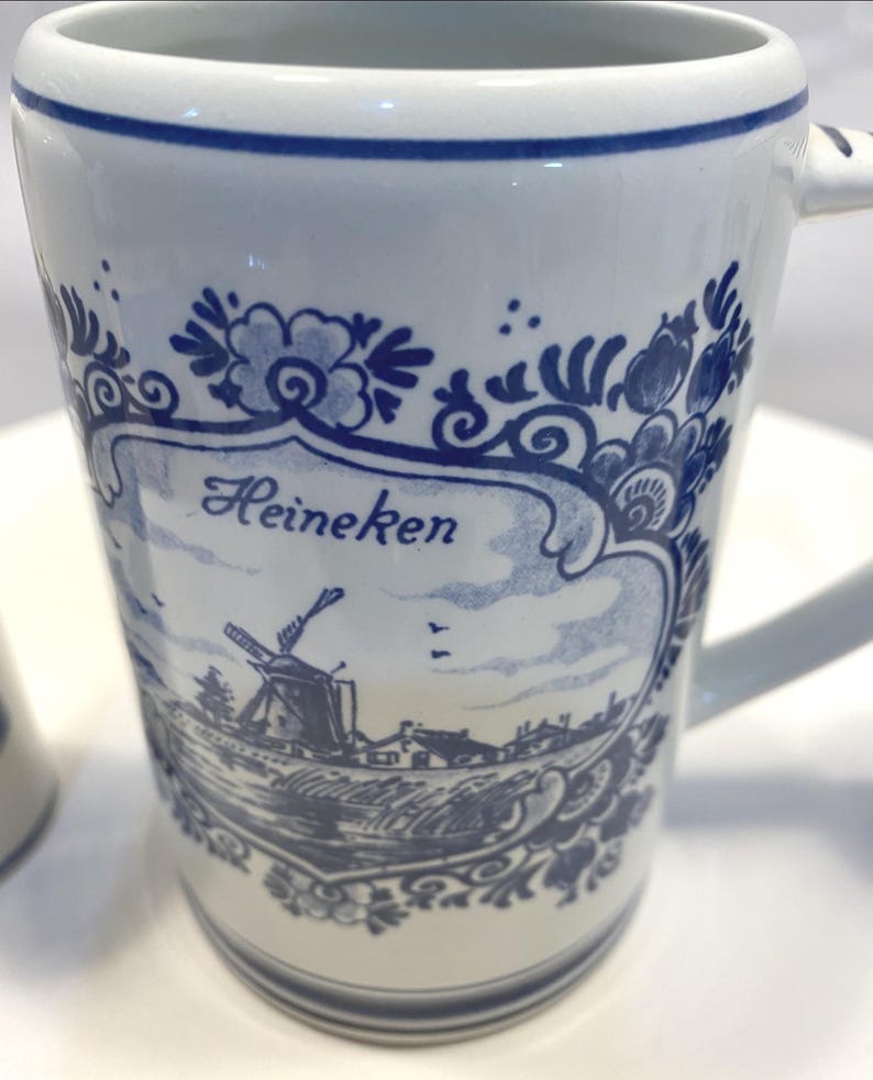 Op de afbeelding: Witte keramische mok met blauw bloemen- en windmolenontwerp. De mok heeft het woord "Heineken" in blauw schrift. Het handvat is wit en de rand heeft een blauwe streep. De mok heeft een klassieke, vintage esthetiek.