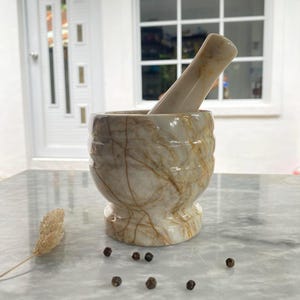 Puede incluir: Un mortero y un pilón de mármol blanco con vetas doradas. El mortero y el pilón están sobre una superficie blanca.