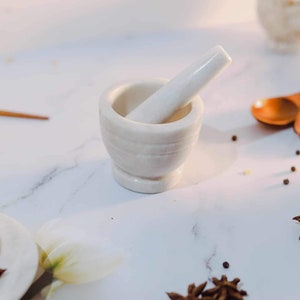 Puede incluir: Conjunto de mortero y maja de mármol blanco sobre una superficie blanca con anís estrellado y granos de pimienta esparcidos alrededor.