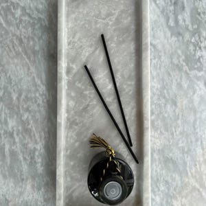 Könnte beinhalten: Ein rechteckiges Tablett aus weißem Marmor mit drei schwarzen Diffusorstäbchen und einer schwarzen Glasflasche mit einem goldenen Quast.