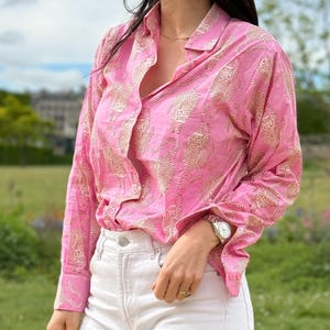 May include: A pink button-down shirt with a white floral pattern. The shirt has long sleeves and a collared neckline. The model is wearing white jeans and a silver watch.