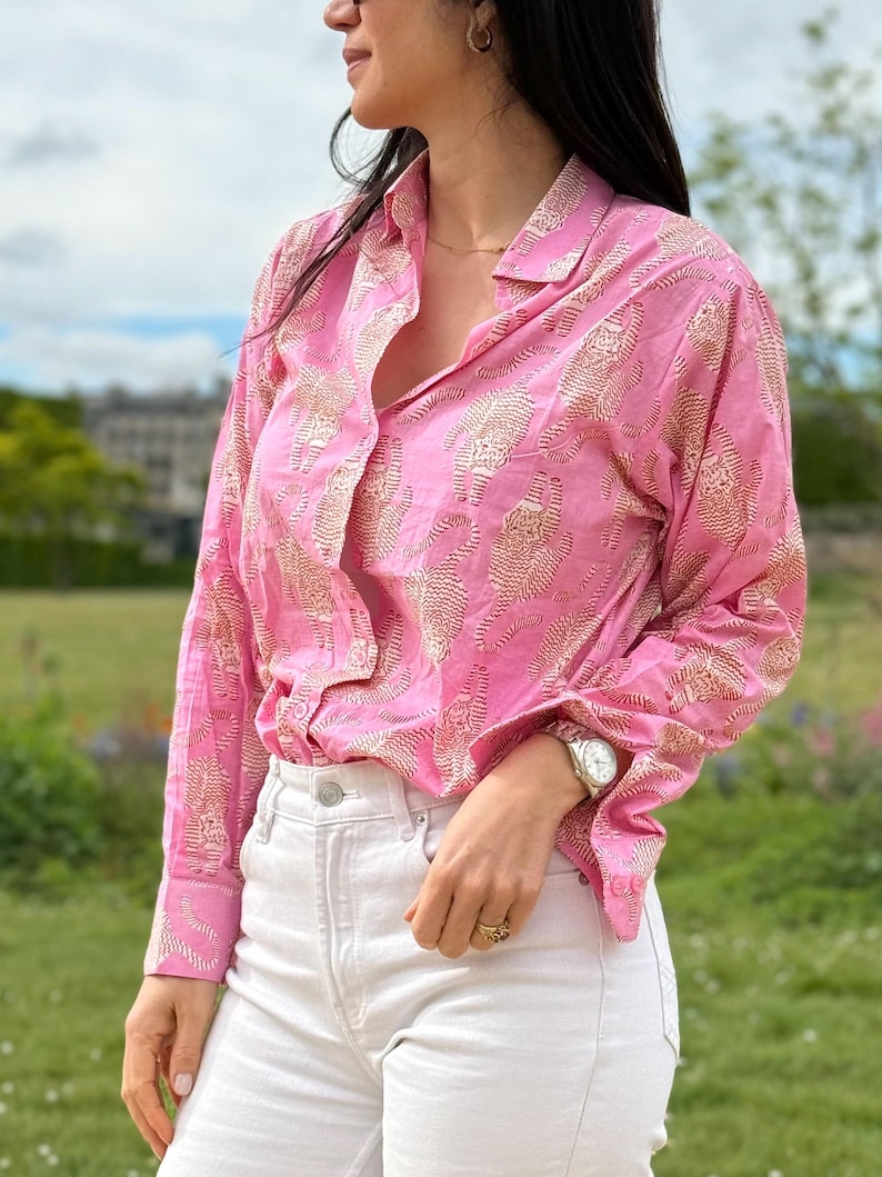 May include: A pink button-down shirt with a white floral pattern. The shirt has long sleeves and a collared neckline. The model is wearing white jeans and a silver watch.
