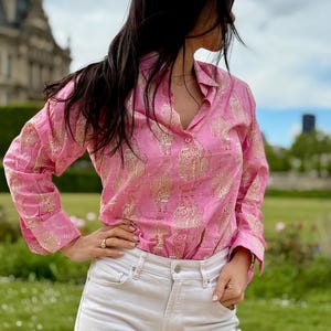 May include: A pink long-sleeved shirt with a gold print of elephants. The shirt has a collared neckline and button closure.