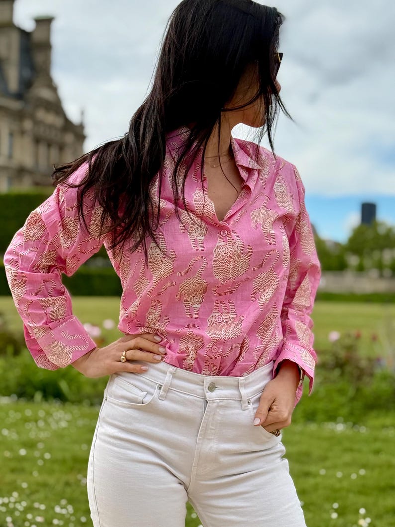 May include: A pink long-sleeved shirt with a gold print of elephants. The shirt has a collared neckline and button closure.
