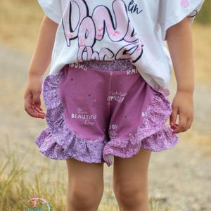 Pode incluir: Uma pessoa jovem vestindo uma camiseta branca com a palavra "Donuts" impressa e shorts roxos com estampa floral branca e rosa. Os shorts têm uma bainha de babado e o texto "It's a Beautiful Day" está impresso na frente.