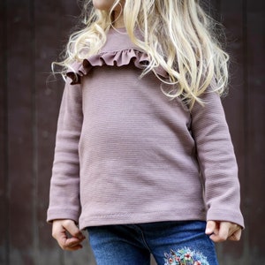 Op de afbeelding: Een mauve lange mouwen wafelgebreide top met een gerimpelde halslijn. De top wordt gecombineerd met een blauwe spijkerbroek. De jeans hebben een bloemenontwerp op de onderbenen. De achtergrond is een donkerbruin houten oppervlak.