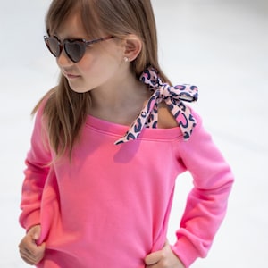 Può includere: Una felpa rosa acceso con maniche lunghe e girocollo. Un fiocco fantasia è legato sulla spalla. Il fiocco ha uno sfondo rosa con una stampa animalier blu scuro. La persona indossa occhiali da sole a forma di cuore.
