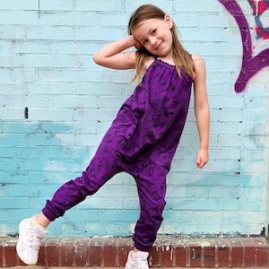 Può includere: Una giovane persona indossa una tuta viola con un motivo stampato nero. La tuta ha una vestibilità ampia e gambe larghe. La persona è in piedi su un gradino di mattoni davanti a un muro di mattoni blu con graffiti viola.